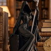 Dunkle, stilisierte Statue einer weiblichen Figur mit einer großen Sense, auf einem Holzuntergrund neben Büchern.