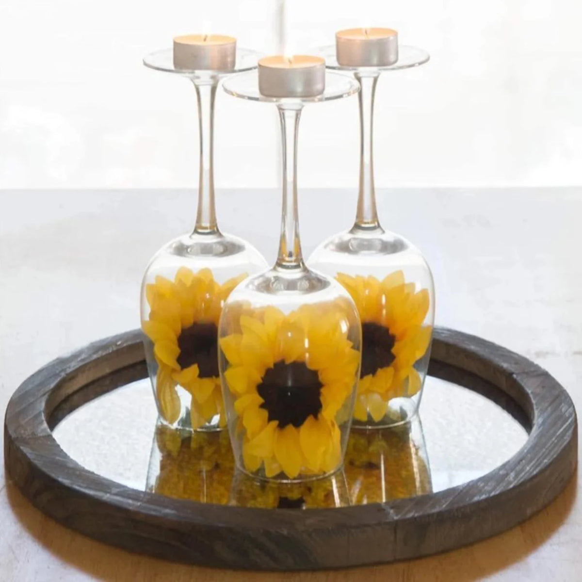 Three glass candle holders with yellow flowers and white tealight candles displayed on a dark wooden tray.