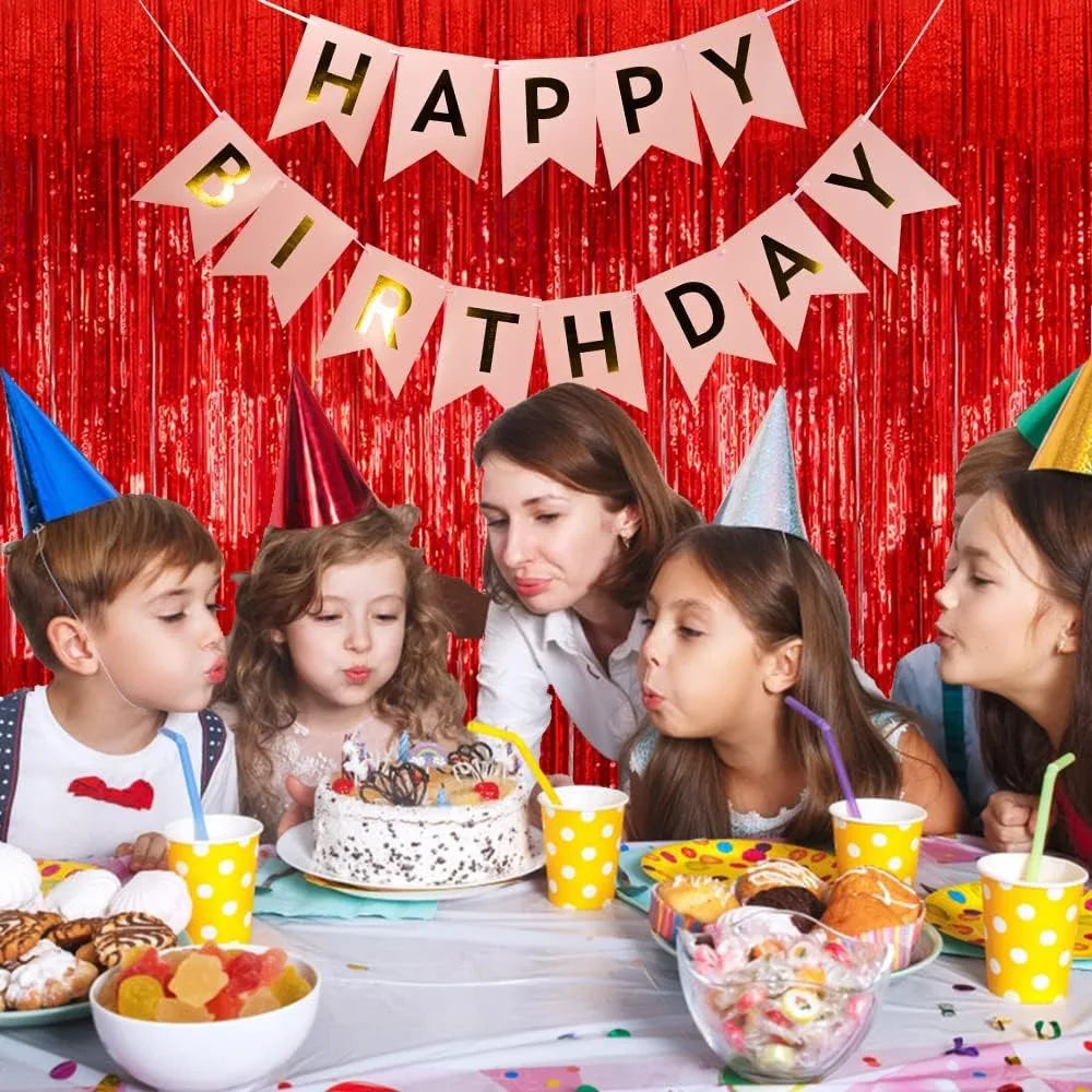 A birthday party setup featuring a red curtain backdrop with a banner, cake, and colorful decorations.