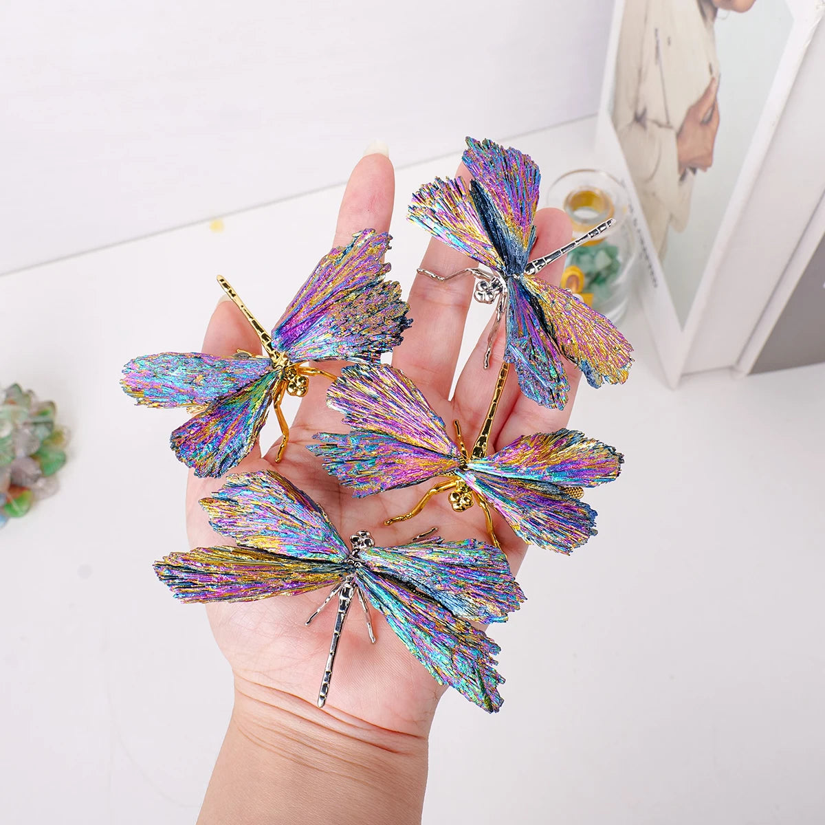 A hand holds several iridescent crystal tourmaline dragonfly figurines.