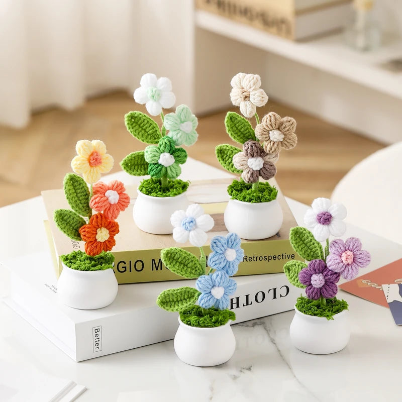 Hand woven crochet flowers in various shades of green and beige are potted in white ceramic cups on a wooden surface.