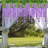 A white floral archway decorated with purple artificial wisteria vine garlands and greenery for an outdoor event.