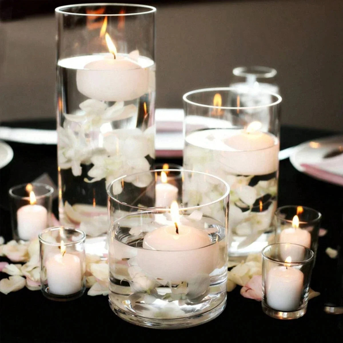 A collection of clear glass candle holders filled with white flowers and lit candles on a dark surface.