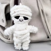 A white mummy skeleton plush keychain is hanging from a strap against a light beige fabric background.