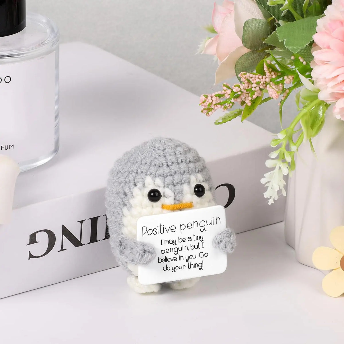 A gray crocheted penguin plush sits next to a white card with an encouraging message on a white surface.