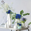 A floral arrangement featuring blue roses and white baby's breath displayed in a white vase on a wooden surface.