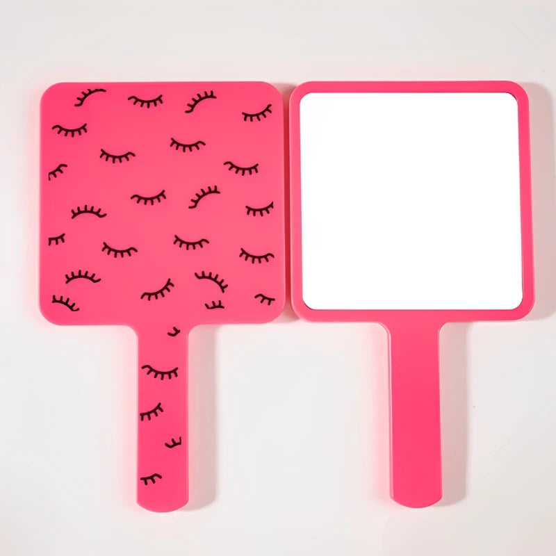 A pink handheld vanity mirror with an ergonomic handle featuring a pattern of black eyelashes.