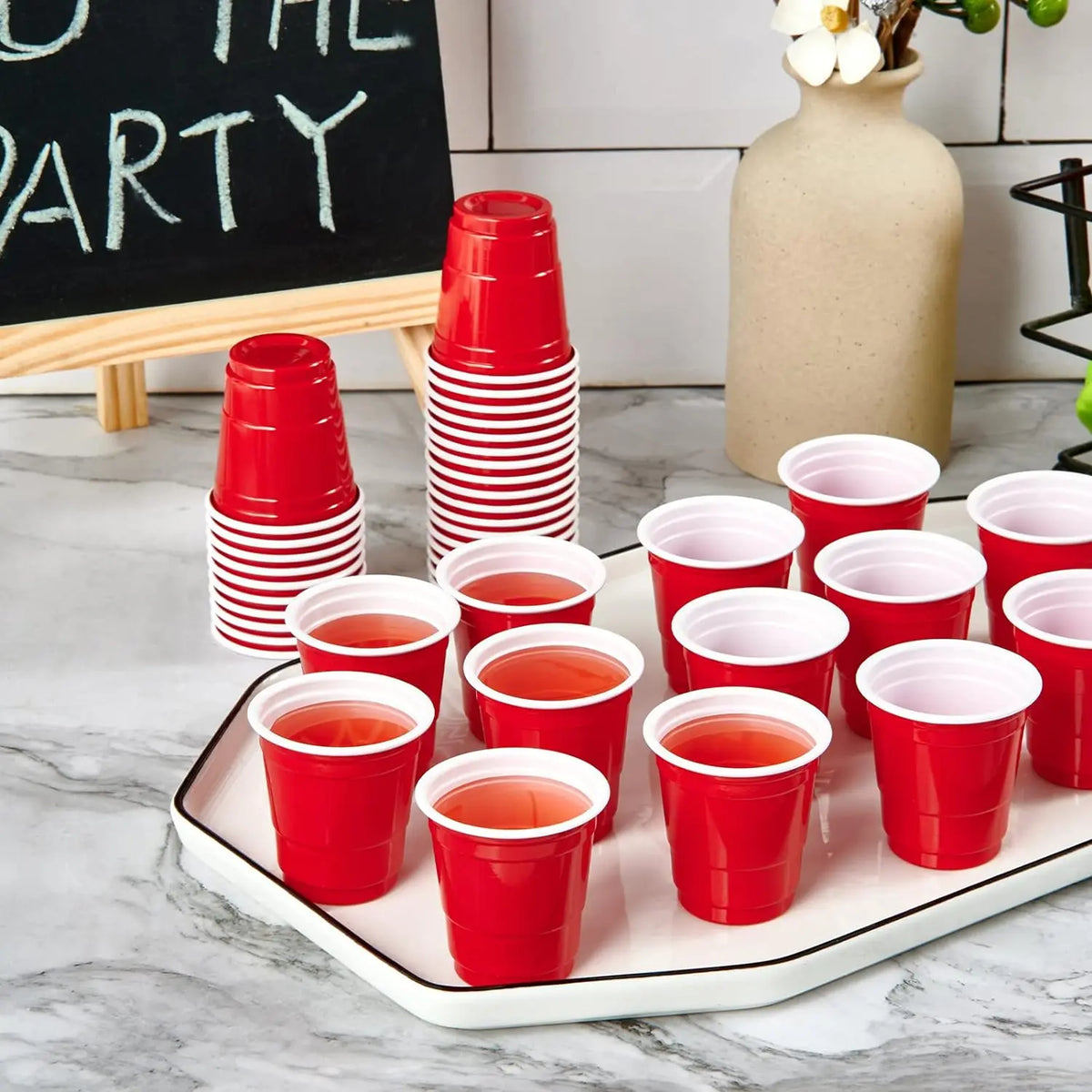 A white platter holds red disposable shot glasses and a stack of red shot glasses on a marble surface.