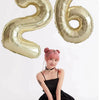 A young woman in a black dress is sitting, surrounded by large champagne gold foil number balloons shaped like 2 and 6.