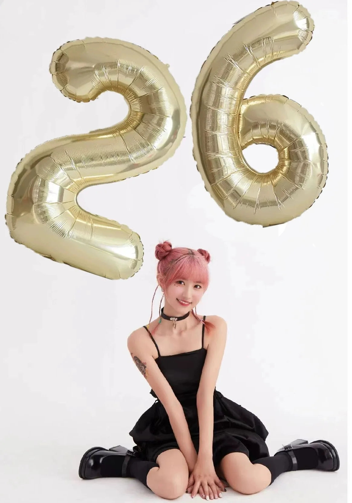 A young woman in a black dress is sitting, surrounded by large champagne gold foil number balloons shaped like 2 and 6.
