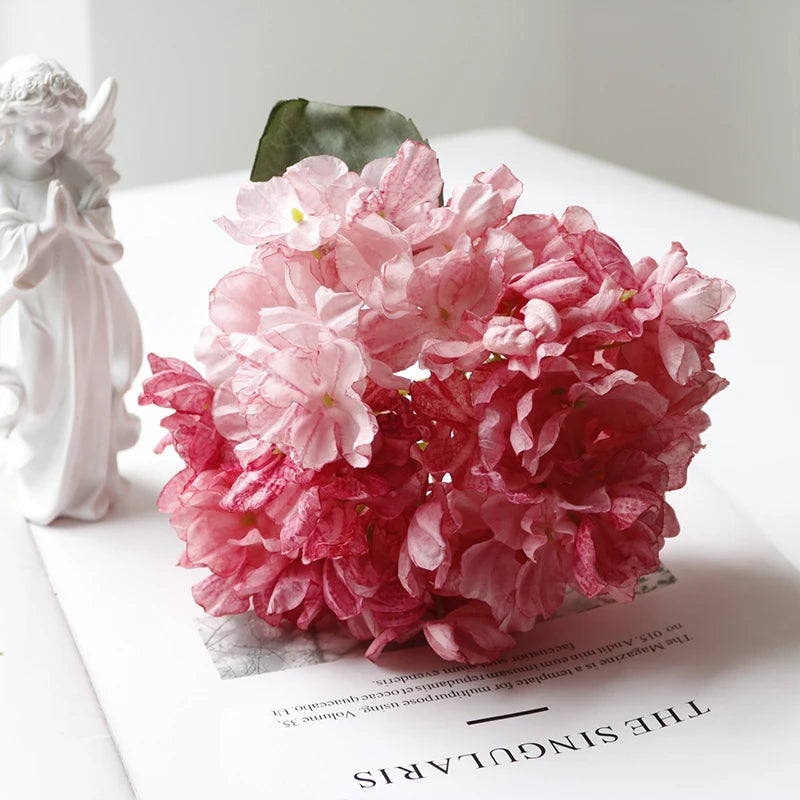 A pink silk hydrangea stem with vintage burnt edge petals displayed next to a white figurine.