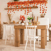 A rustic wooden table set with white chairs and an orange maple leaf garland hanging above.