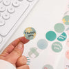 Eine Hand hält einen runden blumigen Dankessticker auf einer weißen Oberfläche.