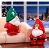 A festive resin Christmas dwarf figurine set featuring a green and white gnome on a pink sofa near a snowy window view.