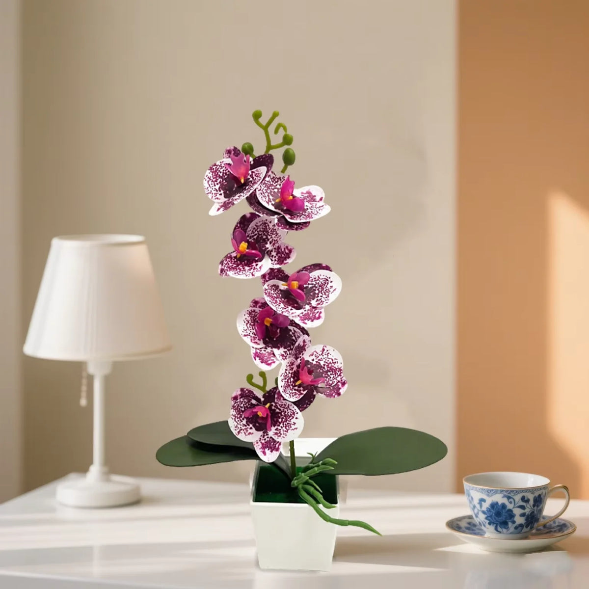 A potted purple orchid plant with green leaves sits next to a white lamp on a table.