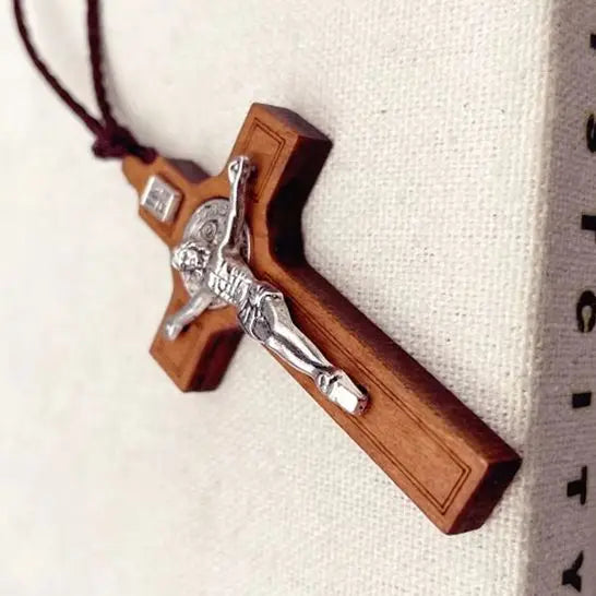 A wooden cross necklace featuring a silver crucifix and a wax rope cord, displayed against a white fabric background.