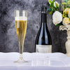 A set of glitter disposable champagne flutes is displayed on a white tablecloth next to a bottle and flowers.