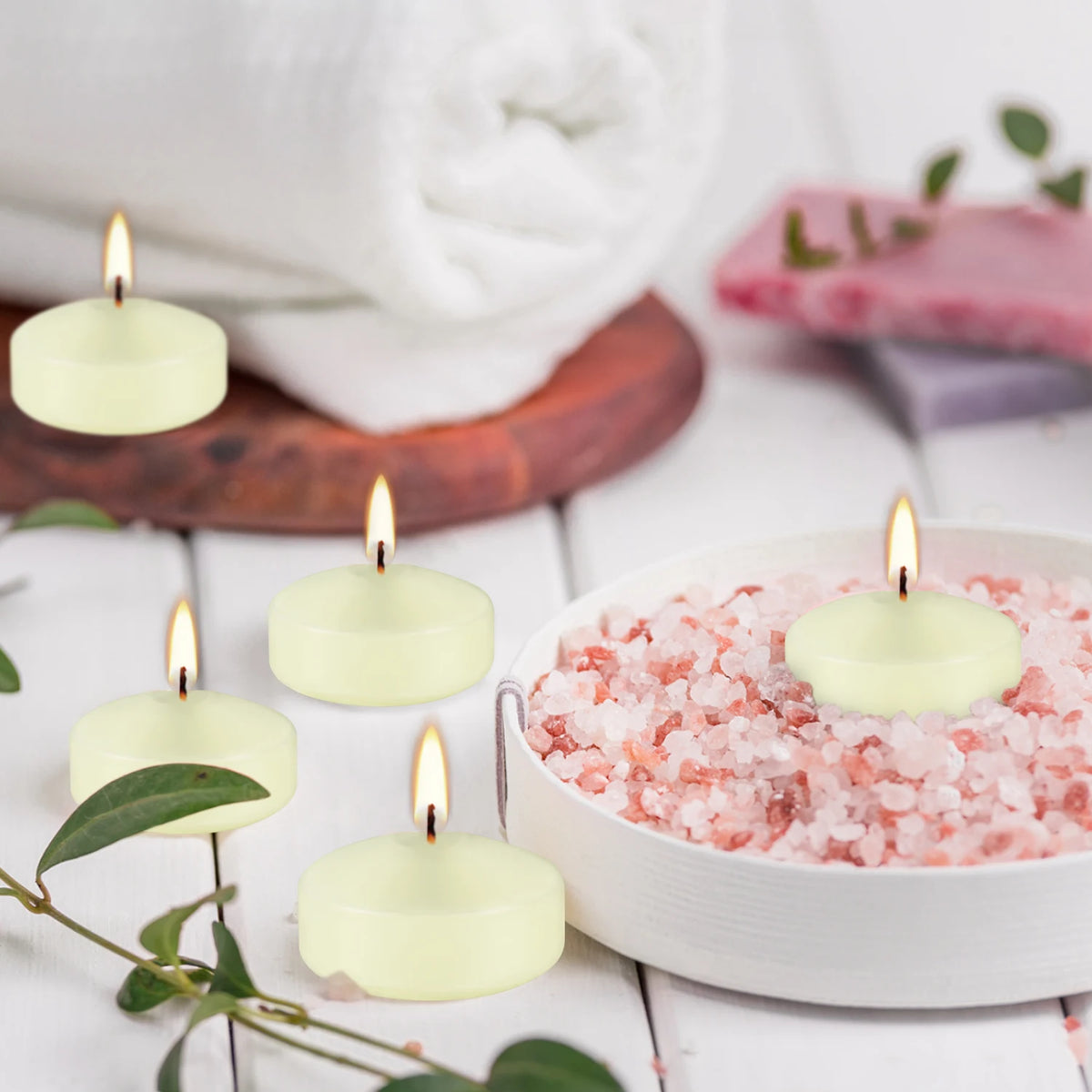 A set of ivory soy wax floating candles in a white bowl surrounded by green leaves and a rolled white towel.
