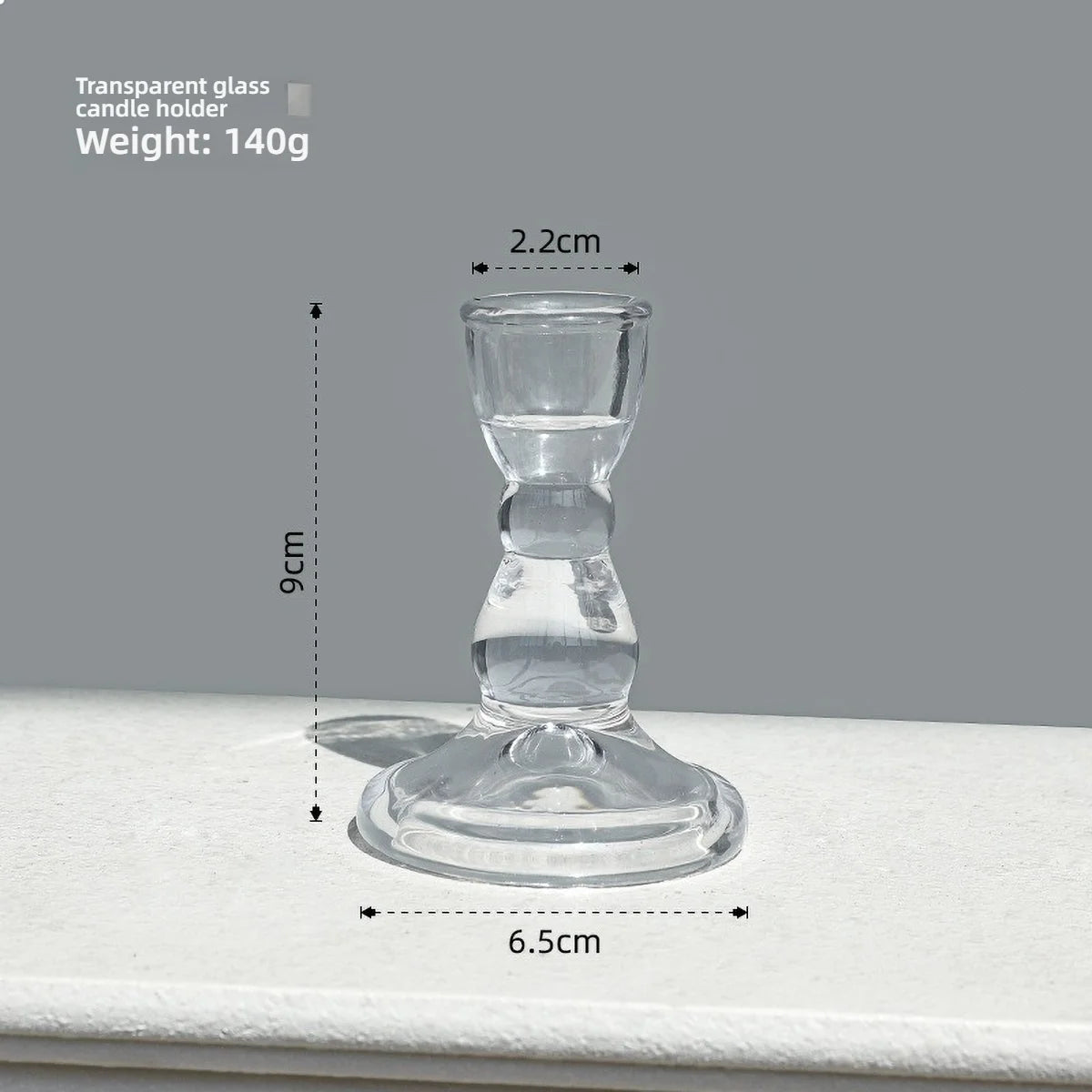 A clear glass pillar candle holder with a wide base and narrow top, shown against a light gray background.