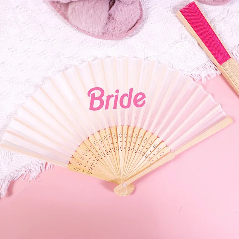 A light pink hand fan with the word Bride written on it, placed on a pink background.