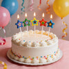 A white birthday cake decorated with colorful star-shaped candles and sprinkles on a pink table.