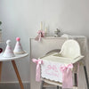A white decorative birthday garland is displayed on a wooden table next to a white baby high chair.
