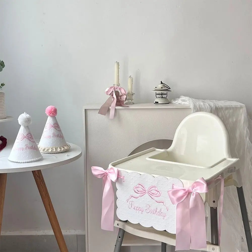 A white decorative birthday garland is displayed on a wooden table next to a white baby high chair.