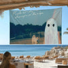 A tapestry featuring Phoebe Bridgers album art, depicting a ghost in the Alps, displayed outdoors near the ocean.