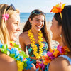 Three women smiling while wearing colorful tropical flower leis and sunglasses on a beach.