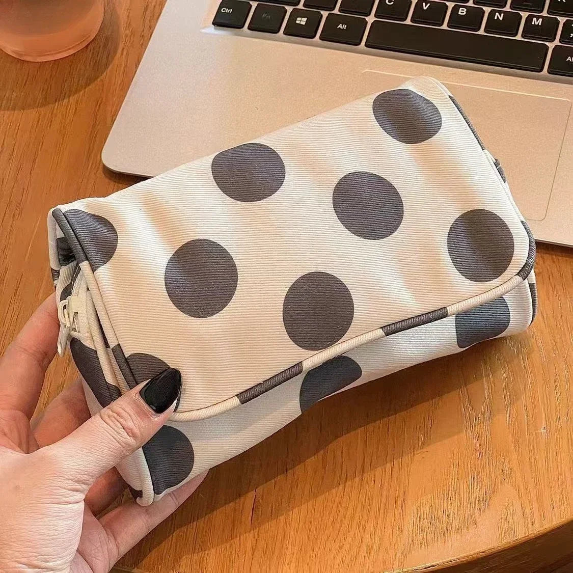 A hand holds a light beige canvas travel makeup bag with dark gray polka dots on a wooden surface next to a laptop.