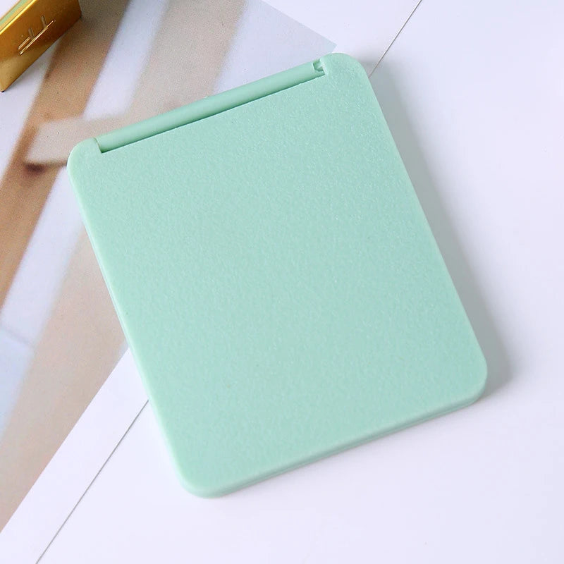A light mint green compact foldable travel makeup mirror with a hinged cover resting on a white surface.
