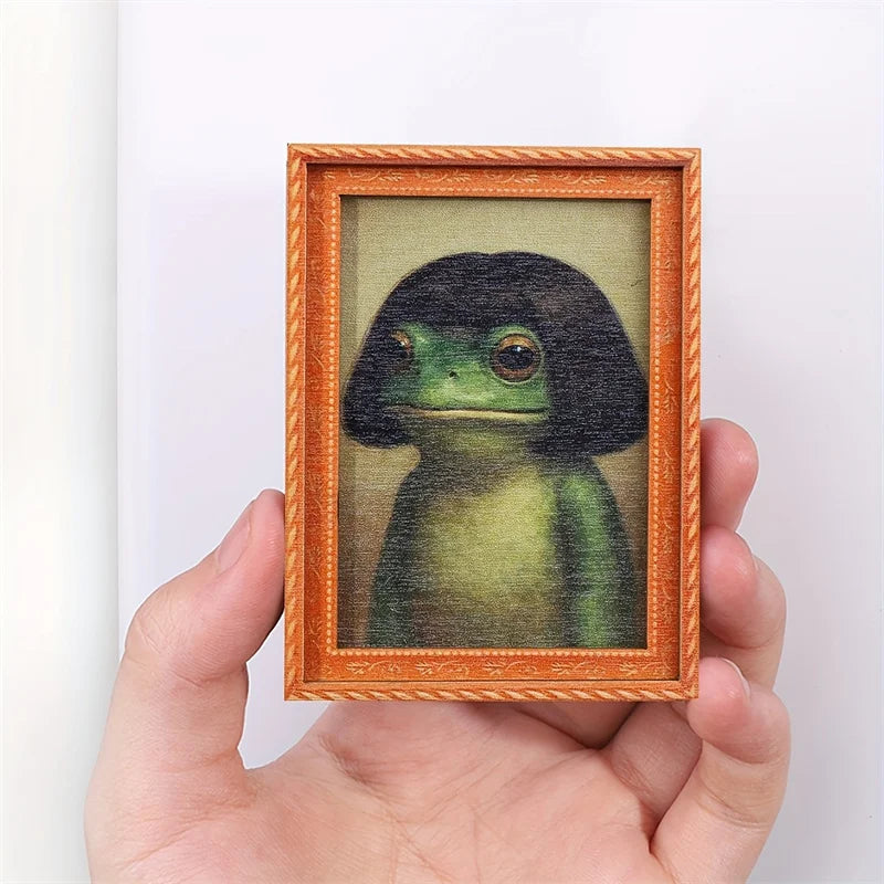 A hand holds a framed watercolor painting of a dark green frog in an orange wooden frame.