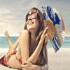 A woman smiling while holding a Japanese foldable hand fan with a Kanagawa wave design on a beach.