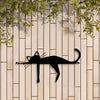 A black silhouette of a cat sculpture is mounted on a light wood panel with green foliage above.