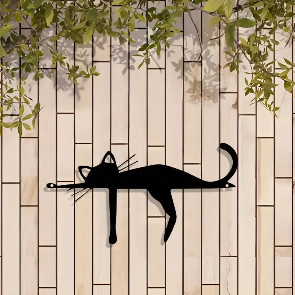 A black silhouette of a cat sculpture is mounted on a light wood panel with green foliage above.