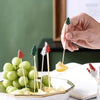 A hand holds a bamboo fruit skewer with a green pine tree decoration over a bowl of green grapes and fruit slices.