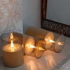 A set of remote control flameless LED candles in a glass holder and cylindrical containers, illuminated with warm light.
