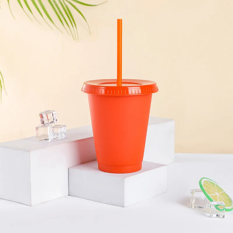 An orange plastic cold cup with a lid and straw is displayed on a white surface next to ice cubes.