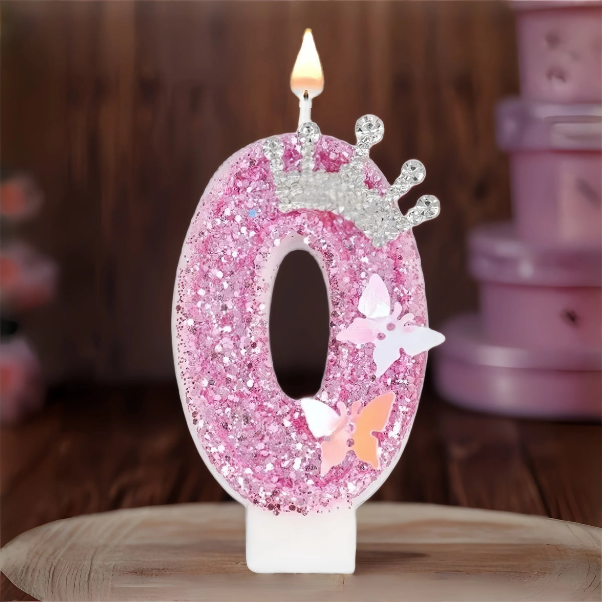 A pink glitter number zero candle with a crown topper and a flame on a wooden surface.