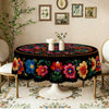 A black embroidered tablecloth with vibrant floral patterns is displayed on a round table in a softly lit room.