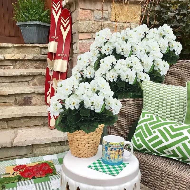 A bouquet of white artificial flowers in a dark green pot sits on a white table next to a mug and pillows.