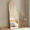 A full length arched wall mirror with a light wood frame reflects a bed and surrounding decor.