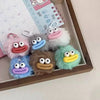 A collection of cute plush sausage-mouth keychains in pink, gray, and blue resting in a wooden tray.