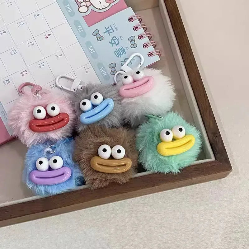 A collection of cute plush sausage-mouth keychains in pink, gray, and blue resting in a wooden tray.