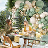 A woman in a yellow top is surrounded by a large balloon arch featuring sage green, gold, and white balloons.