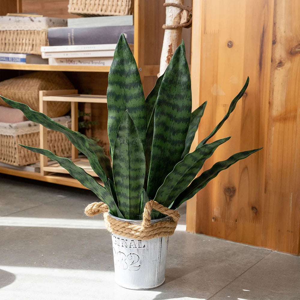 Una alta planta de serpiente artificial en una maceta blanca con cuerda natural, exhibida en un estante de madera.