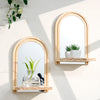 A set of geometric wooden wall shelves holding a glass vase with green stems and a small potted plant in an arched mirror fra