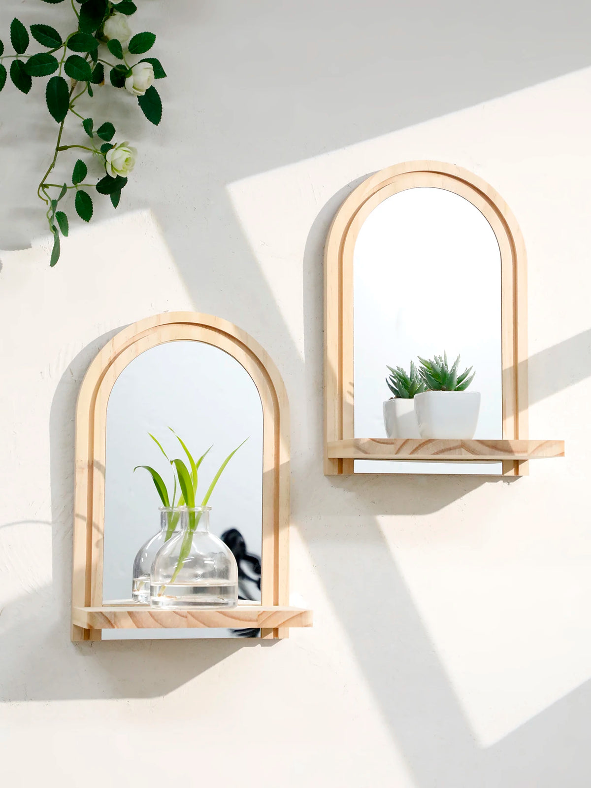A set of geometric wooden wall shelves holding a glass vase with green stems and a small potted plant in an arched mirror fra