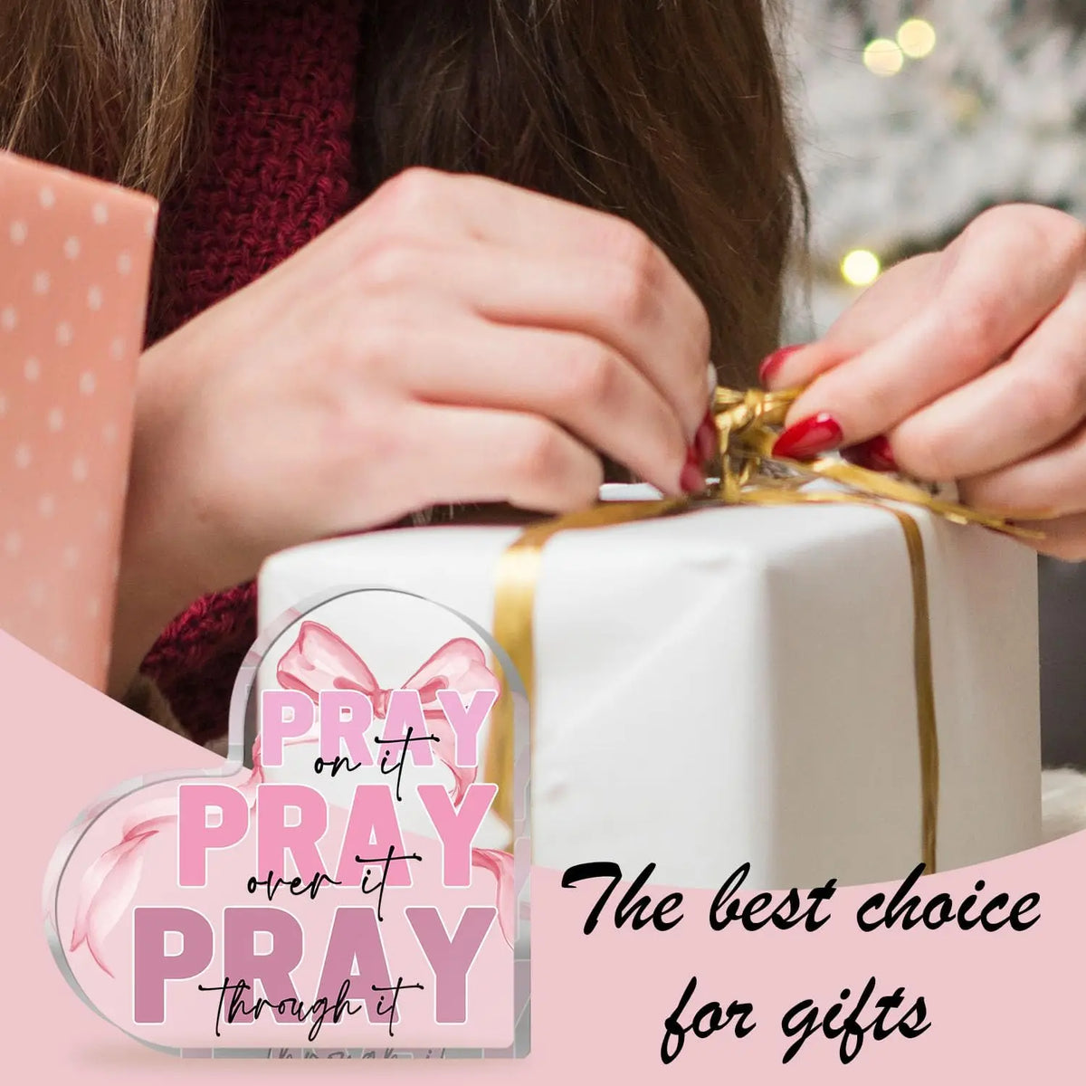 A white gift box tied with a gold ribbon is being held by hands, with a pink acrylic heart sign featuring the text Pray on it