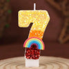 A candle shaped like the number 7 made of yellow sequins, topped with a rainbow and red glitter.